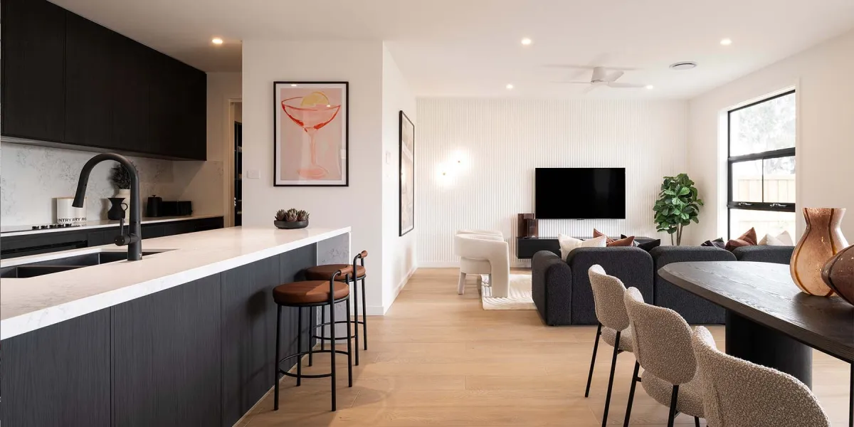 Modern open-plan living room and kitchen in the Coolum 302 display home, featuring a black and white kitchen with marble countertops on the left, bar seating with brown leather stools, light wood flooring throughout, a dark upholstered sofa with throw pillows in the center, a wall-mounted flat-screen TV, white vertical wall paneling, potted plant, framed cocktail artwork, dining table with beige chairs on the right,