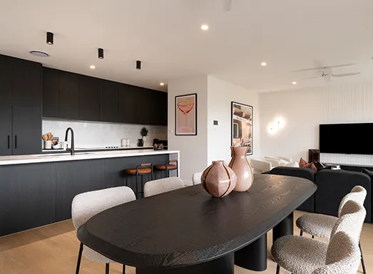 Modern open-plan apartment interior featuring a black kitchen with white countertop on the left, a black oval dining table in the foreground with decorative pottery, comfortable seating areas with cream-colored chairs, wall art, and a television in the background, exemplifying contemporary living space design in Coolum.